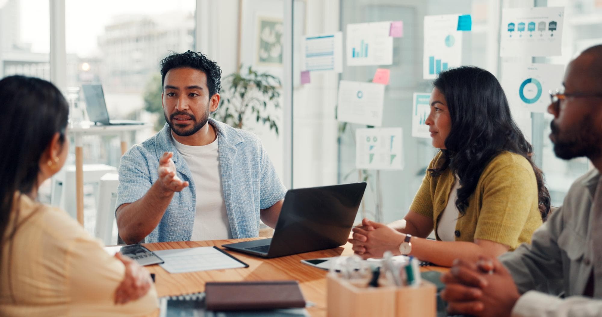 Team collaborating in a bright conference room—discussion around a wooden table with notes and visuals on glass behind them.
